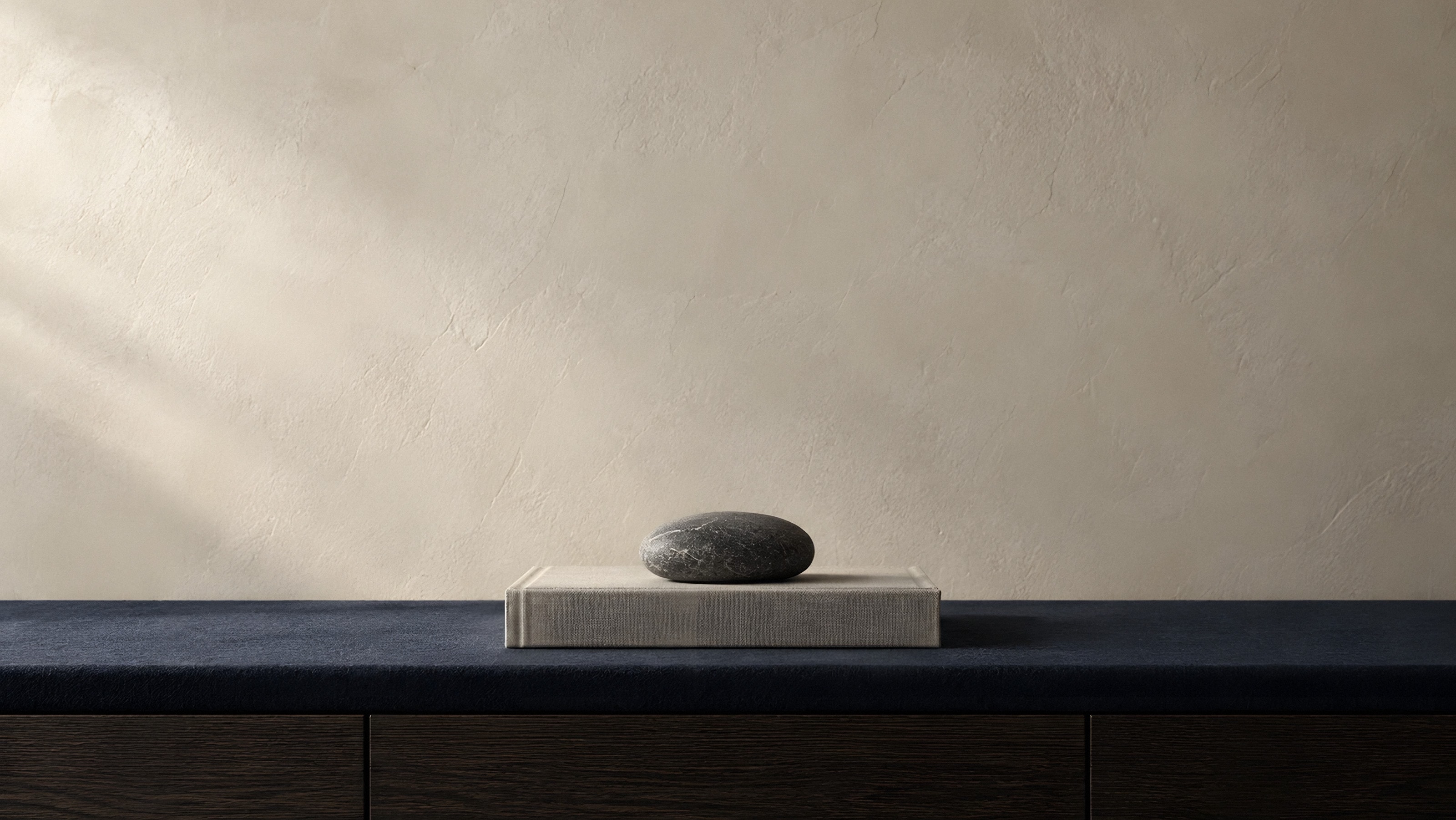 A grey cloth book and a river stone on a navy console against a plaster wall